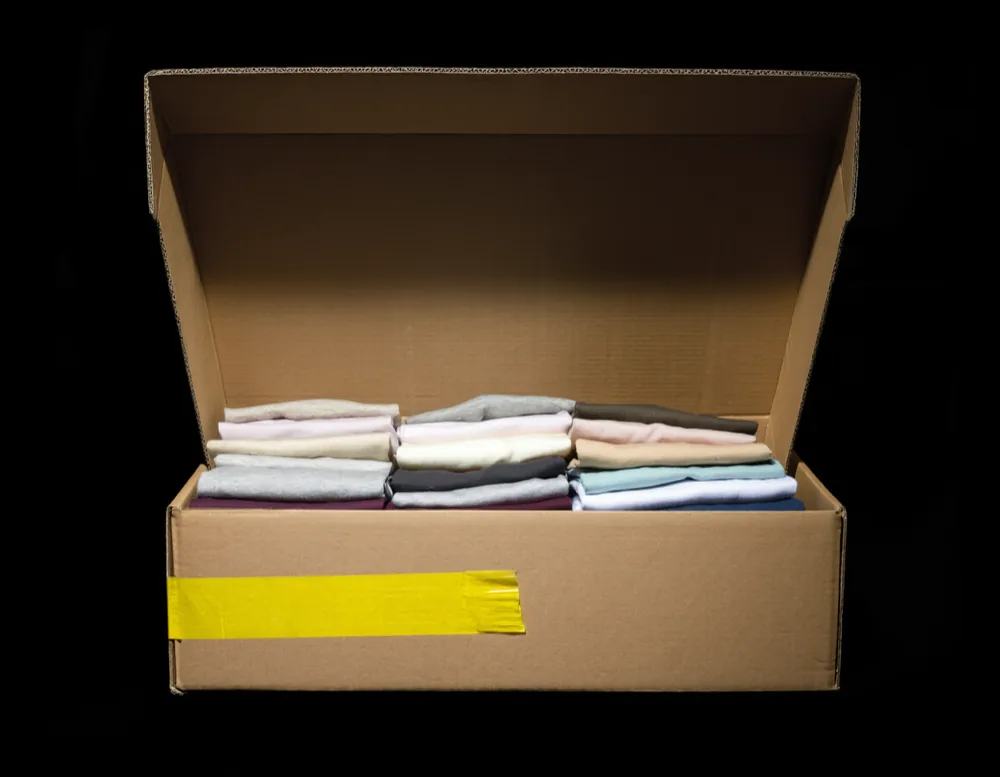 An open cardboard crate packed with folded cotton t-shirts, a strip of yellow tape across the front edge.