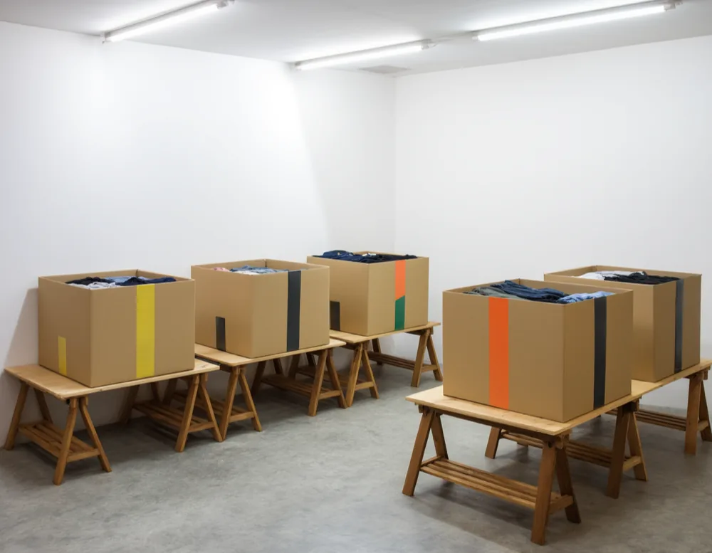 Open cardboard crates of folded clothes on wooden trestle tables, each crate marked with a coloured tape stripe.